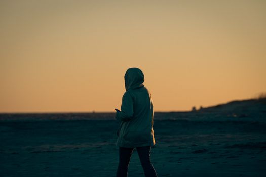 Person in a hooded jacket strolling along a beach at sunset, evoking solitude and tranquility.