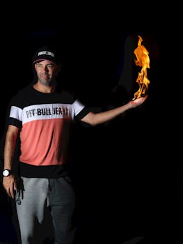 A man in casual attire holds a flame in his hand against a dark background.