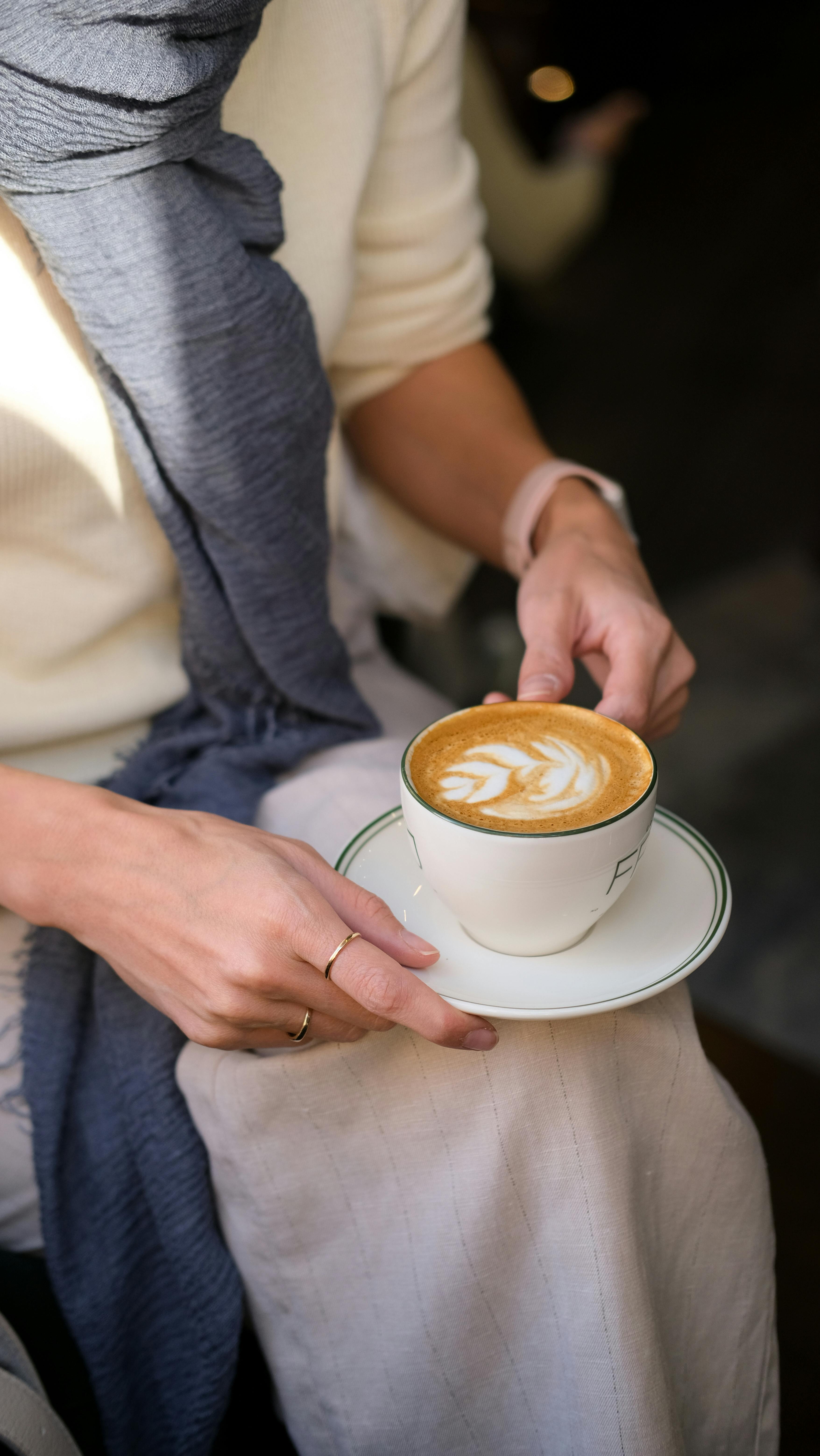 Warm latte with latte art held by woman in cozy setting, wearing scarf.