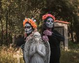 Two women in Dia de los Muertos makeup with floral crowns pose by an angel statue.