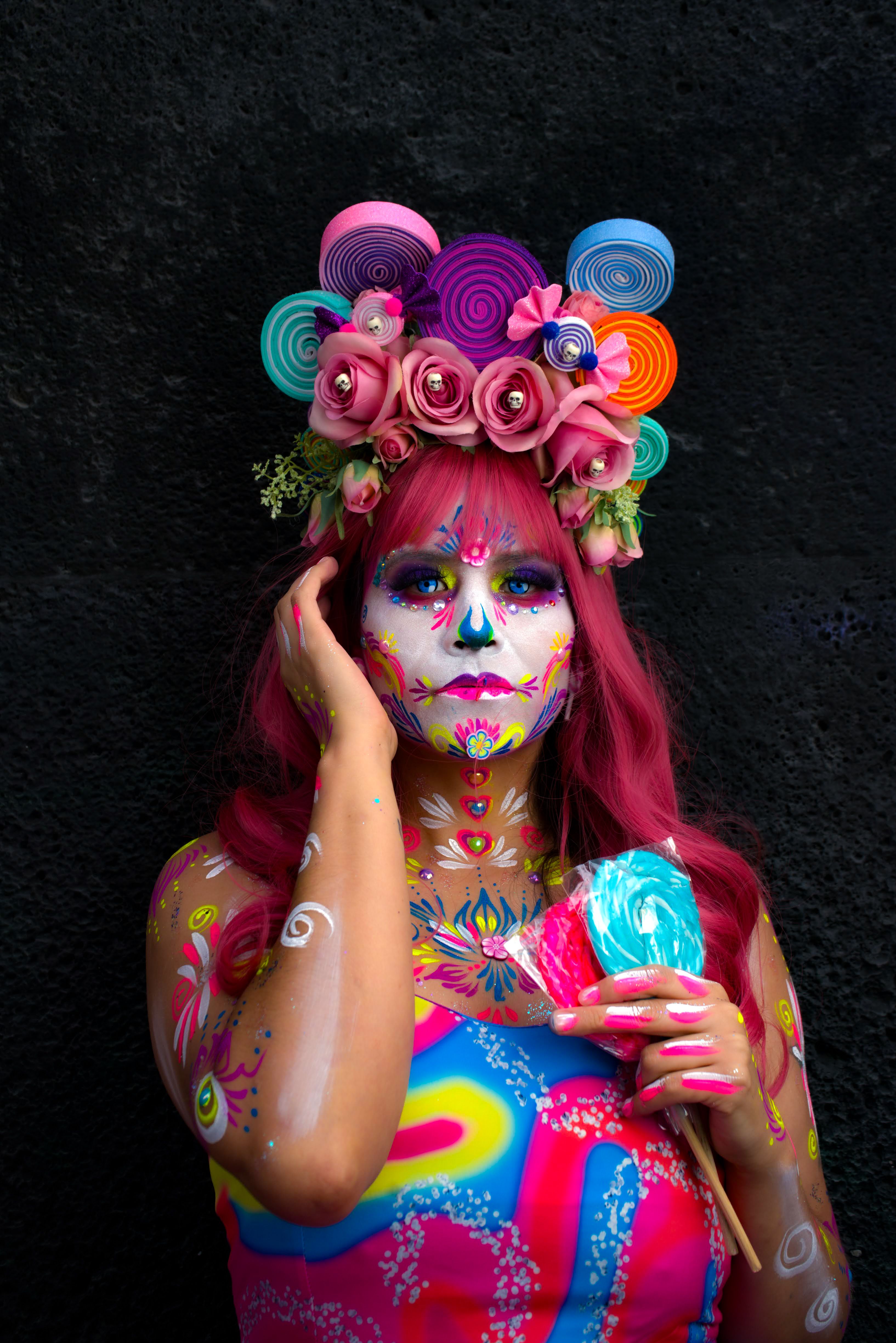 A colorful and artistic portrait featuring a woman with face paint and a decorative headpiece, holding candy.