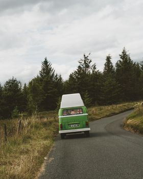 A vintage green van drives on a scenic winding road through a forest.