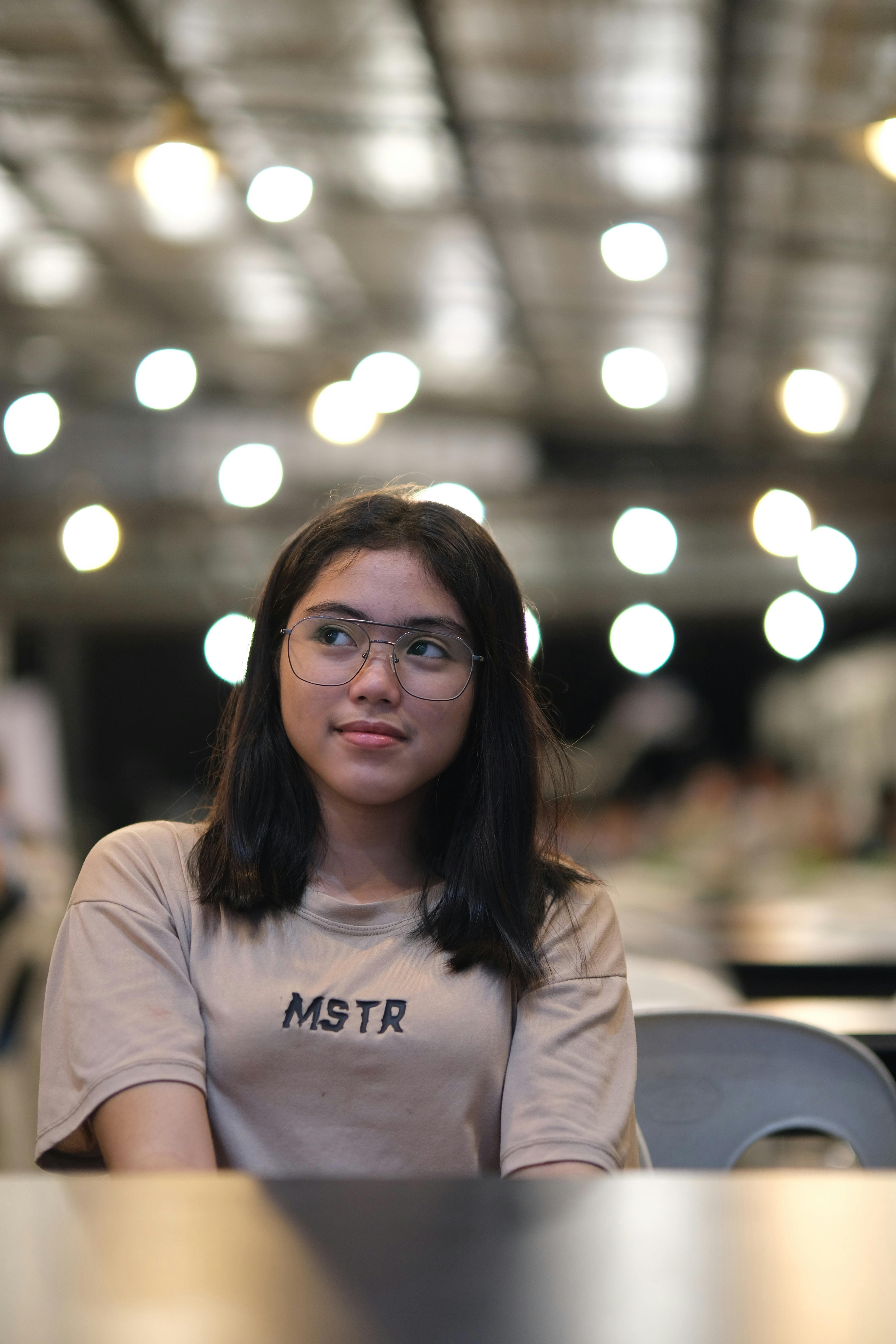 Young woman with glasses in a casual indoor setting with blurred lights.