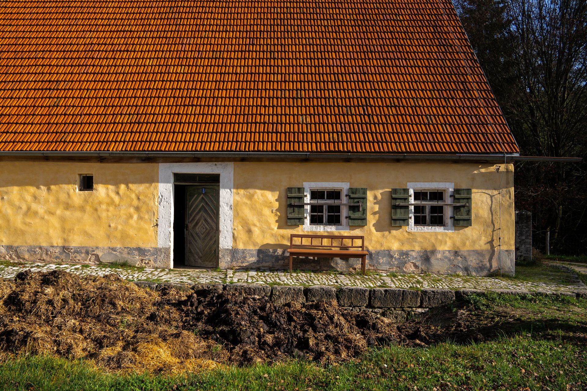 A picturesque yellow farmhouse with an orange tile roof surrounded by autumn foliage and natural landscape.