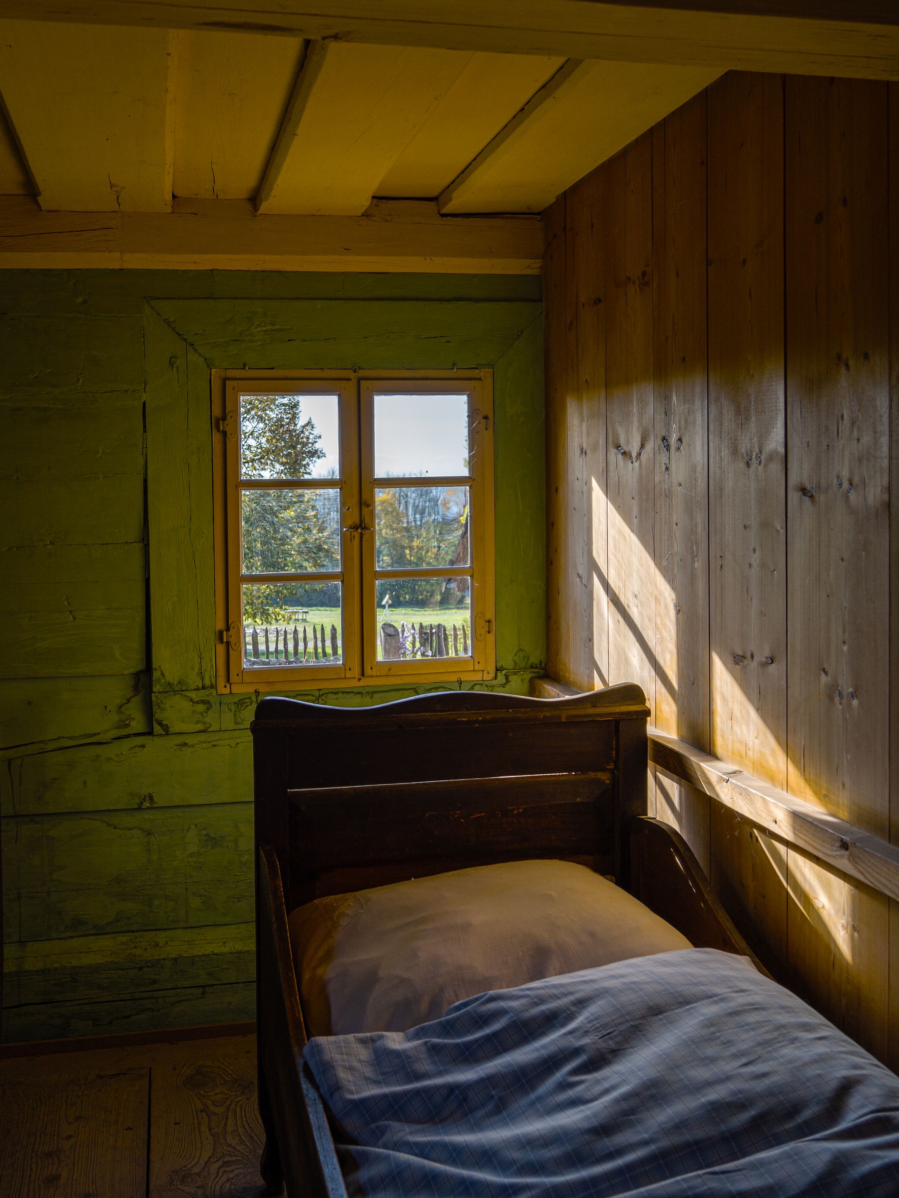Sunlit farmhouse bedroom with timber walls, a simple bed, warm wood furniture and a rural view through the window