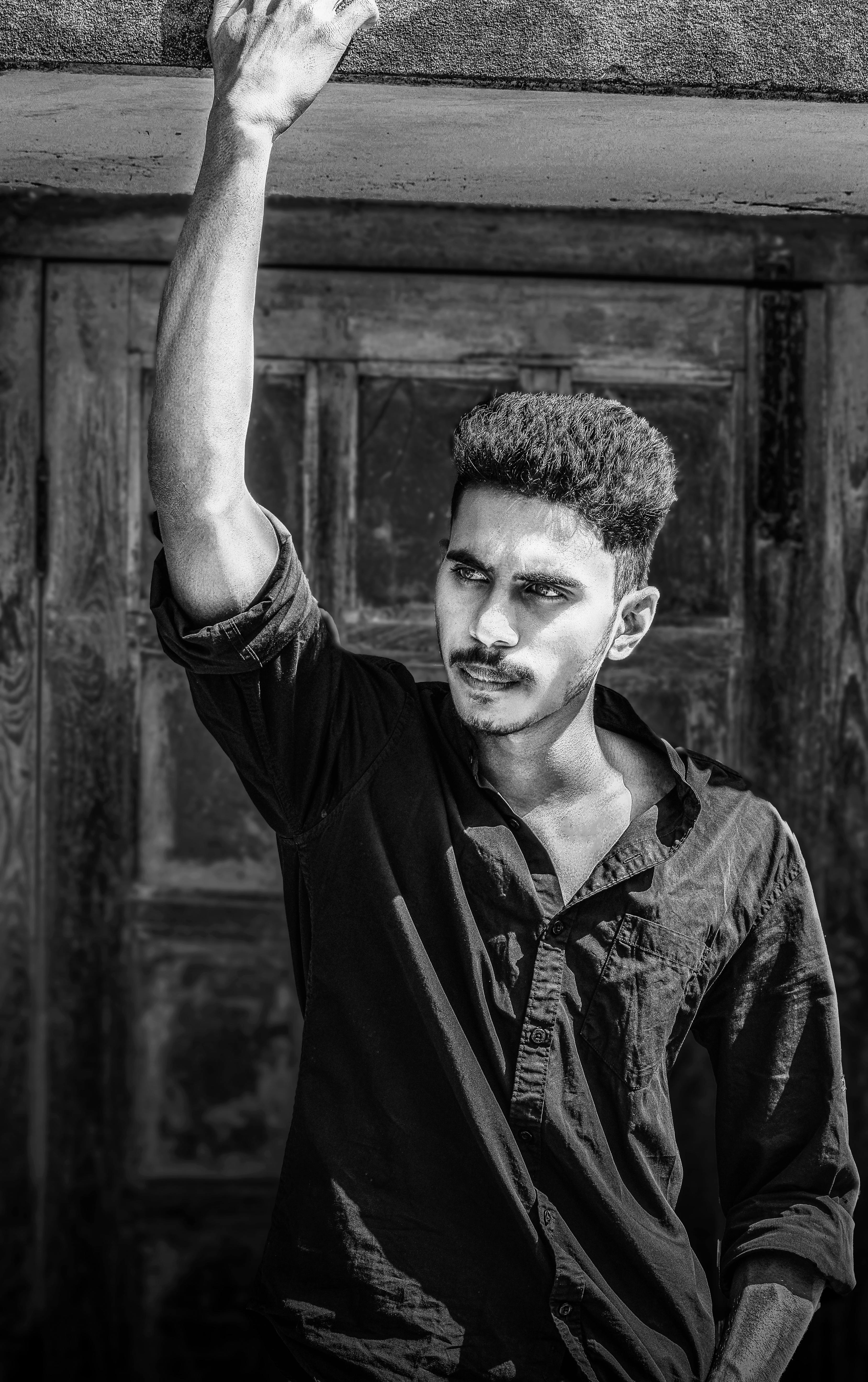 A black and white portrait of a young man standing in front of a wooden door, looking contemplative.