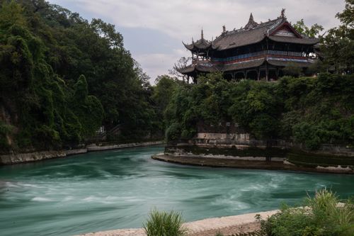 Free A historic Chinese pavilion surrounded by lush greenery by a vibrant river bend under cloudy skies. Stock Photo