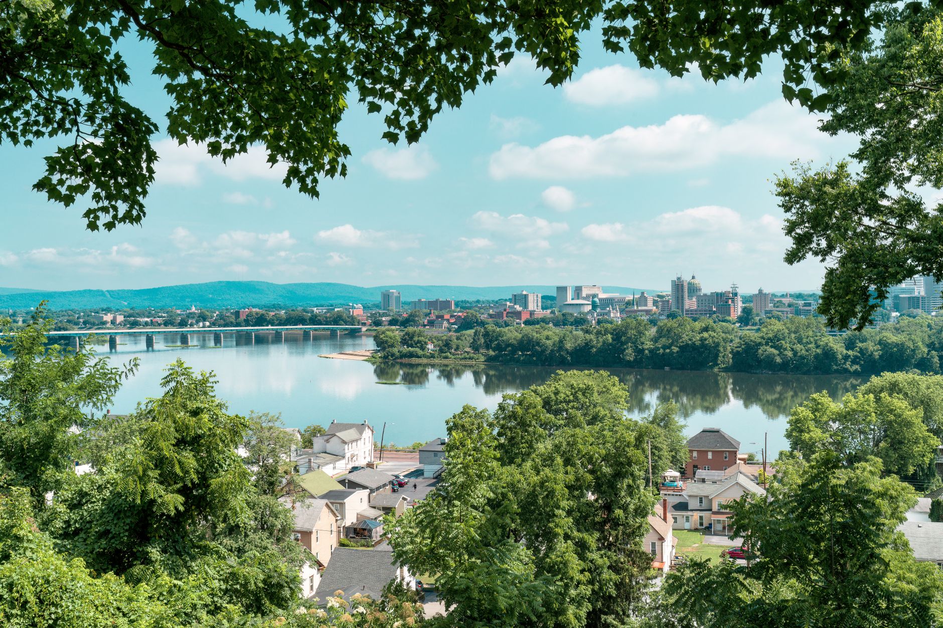 A picturesque view of a city skyline reflected in a calm river, framed by lush trees.