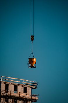 A yellow concrete bucket suspended by a crane at a construction site against a clear sky.