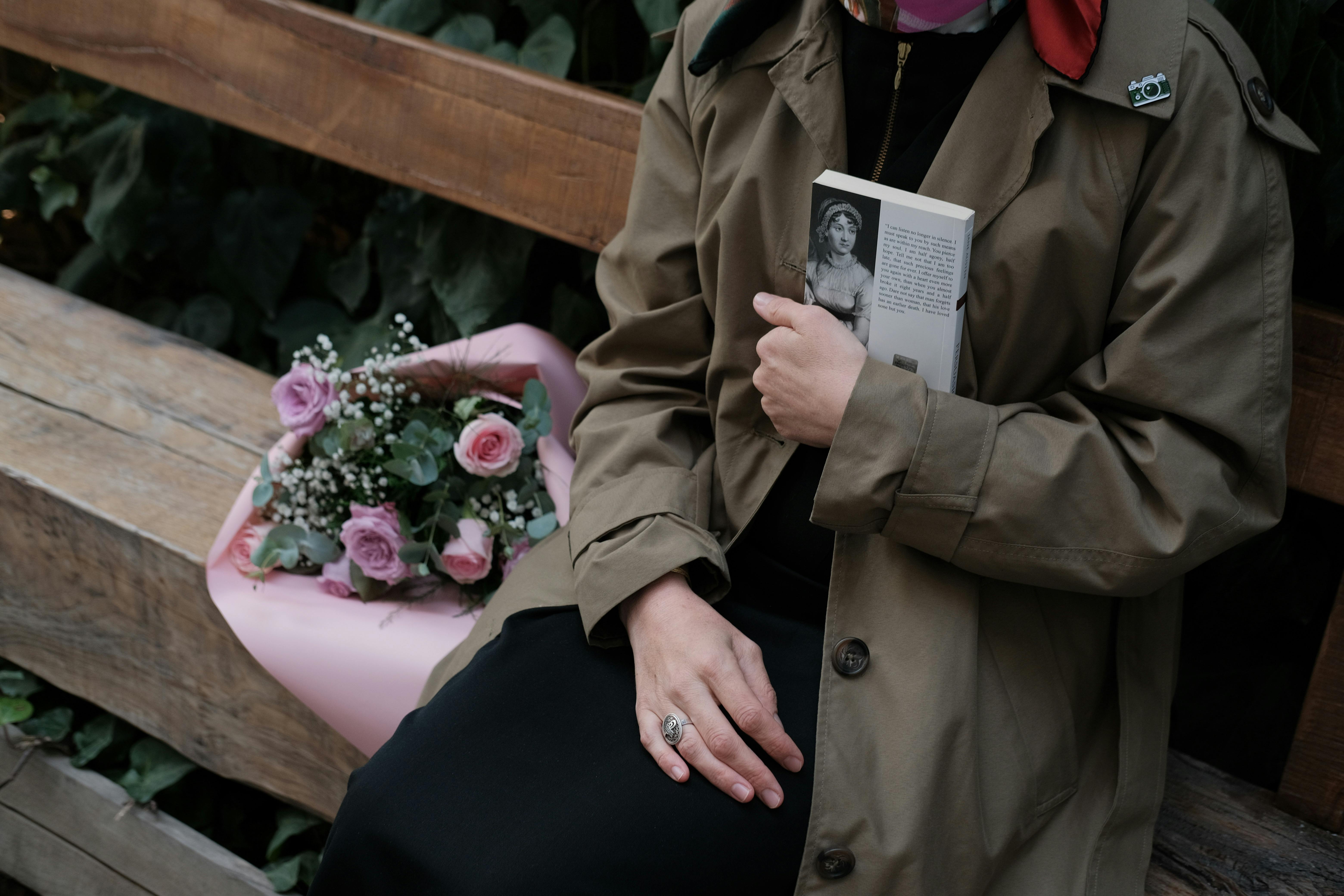 무료 코트를 입은 사람이 나무 벤치 위에 책과 꽃을 놓고 있다. 스톡 사진