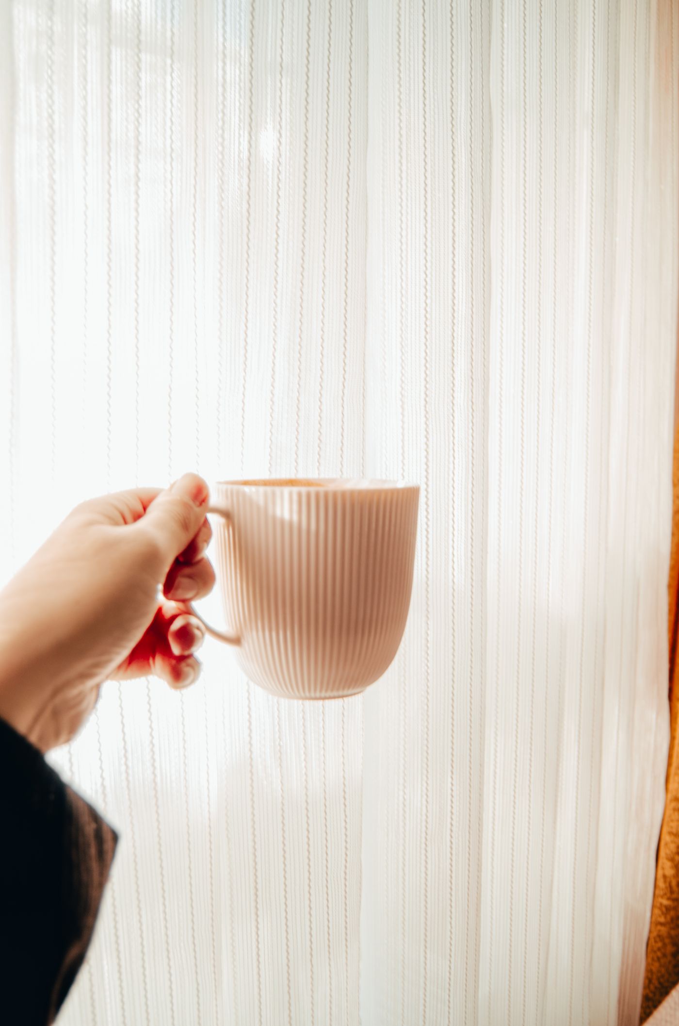 Hand holding a warm mug by a sunlit sheer curtain