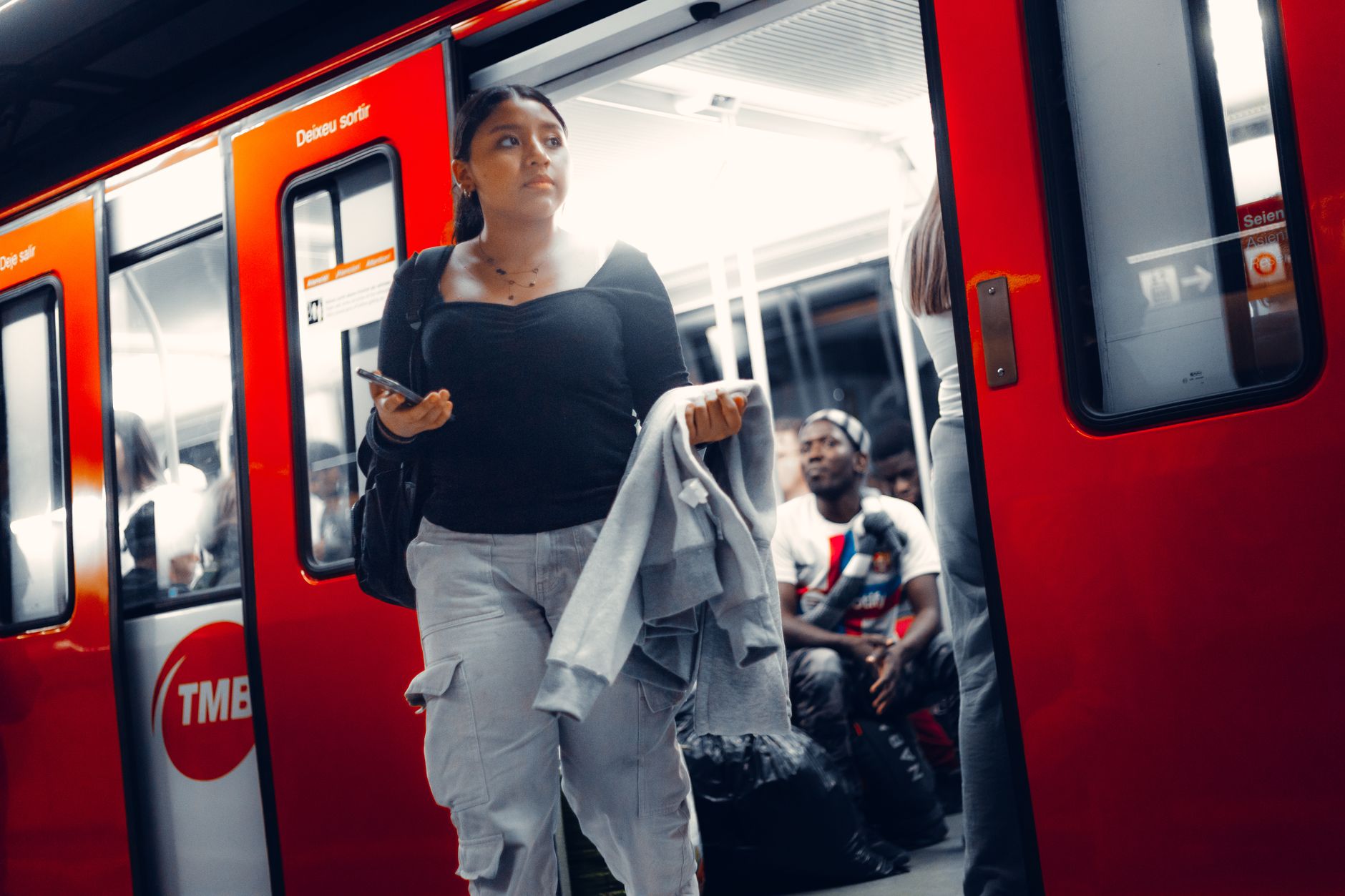 Woman exits vivid red train on busy city metro platform holding smartphone.