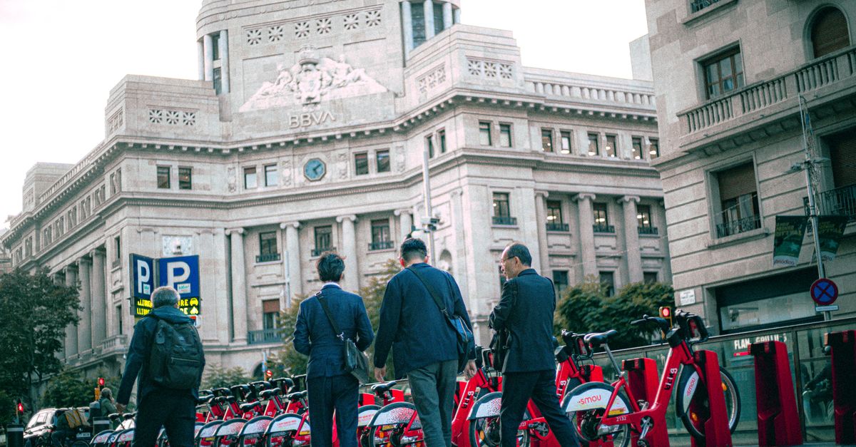 Hombres de negocios caminan frente al edificio del BBVA en la ciudad con bicicletas de alquiler.