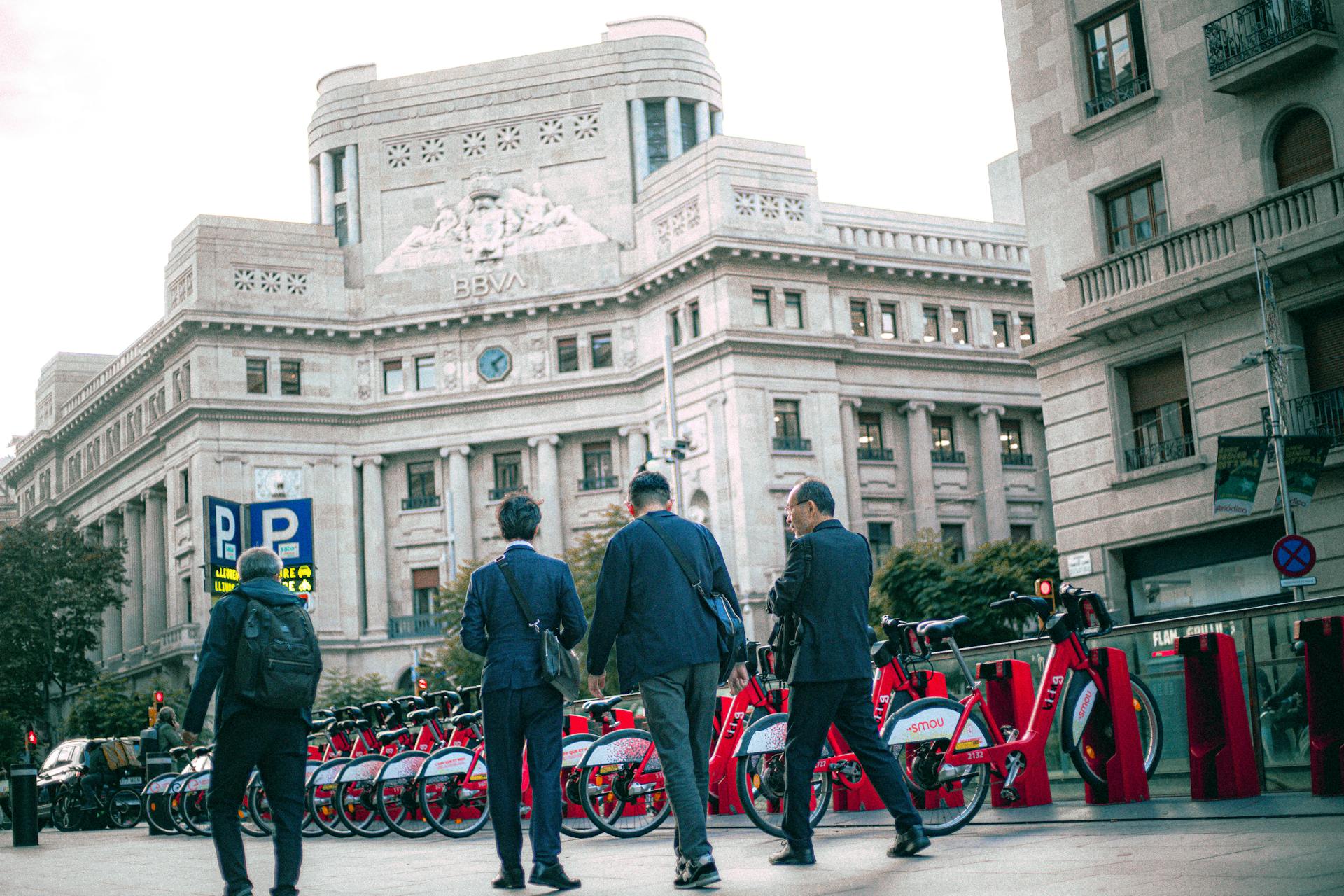 Businessmen walk past BBVA building in city with rental bikes.