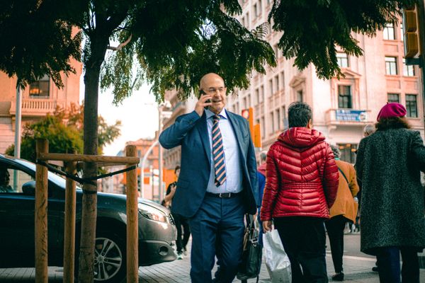 Businessman in a blue suit walking on a bustling city street while talking on his phone.