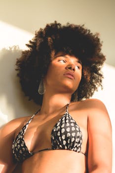 一位留着自然头发、穿着比基尼上衣的时尚女性，若有所思地对着白色墙壁摆出姿势