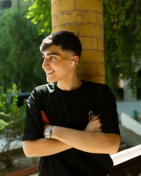 Smiling young man with earbuds leaning against a brick wall outdoors.
