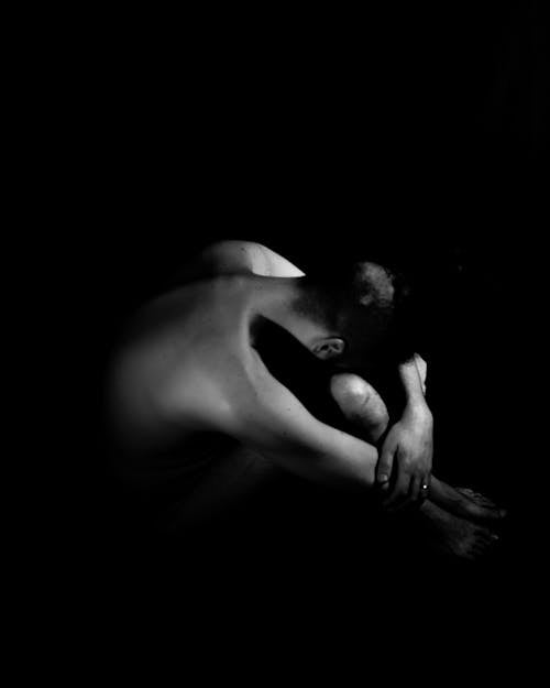 Fotografia Em Tons De Cinza De Uma Pessoa Nua Sentada No Chão Com A Cabeça Inclinada Sobre Os Joelhos Em Um Lugar Escuro