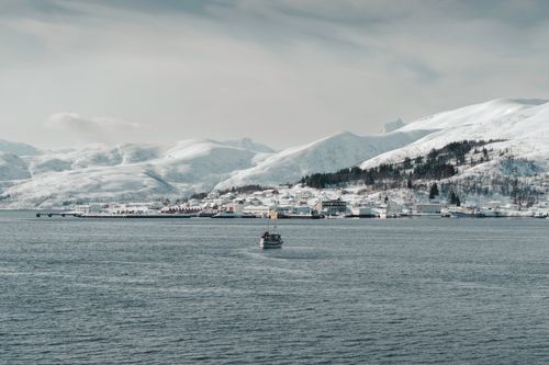 Free A peaceful winter landscape featuring a boat on a Norwegian fjord with snow-covered mountains and shoreline houses. Stock Photo