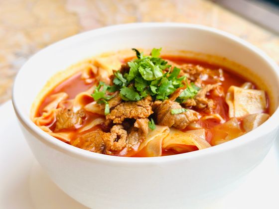White bowl of wide flat noodles in red tomato broth with tender beef slices and cilantro