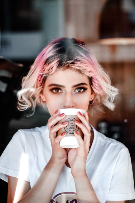 Portrait of a young woman with pink hair holding a cup in a cafe setting.