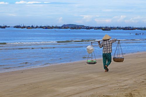 Free A fisherman carries goods along a sandy beach with fishing boats in the background. Stock Photo