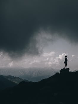 Silhouette of a lone person standing on a mountain peak under dramatic, dark clouds.