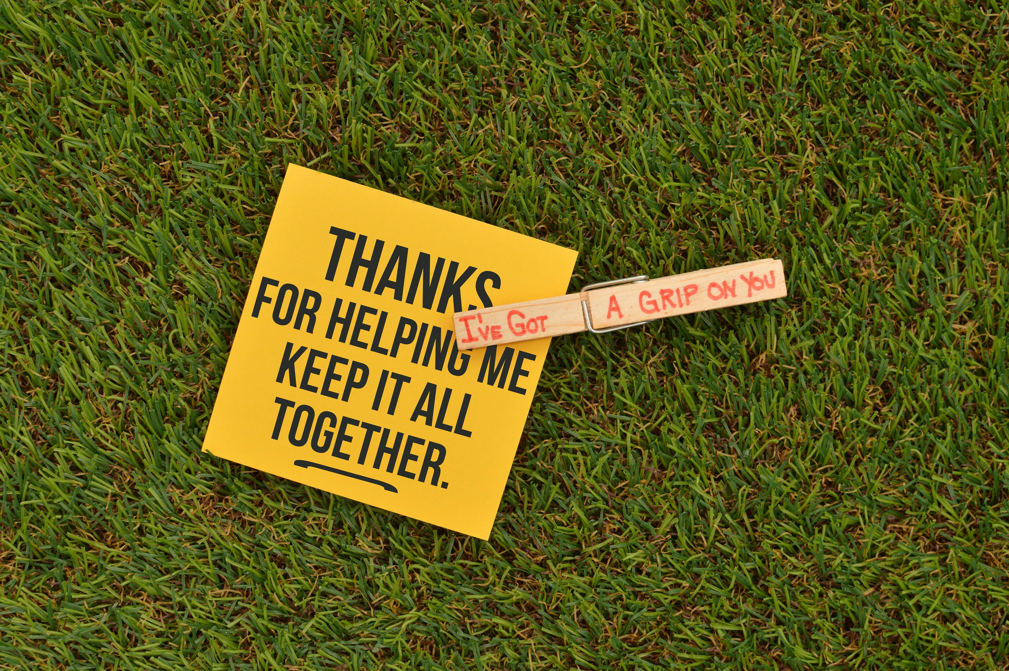 A yellow note with a message of thanks on vibrant green grass, held by a clothes pin.