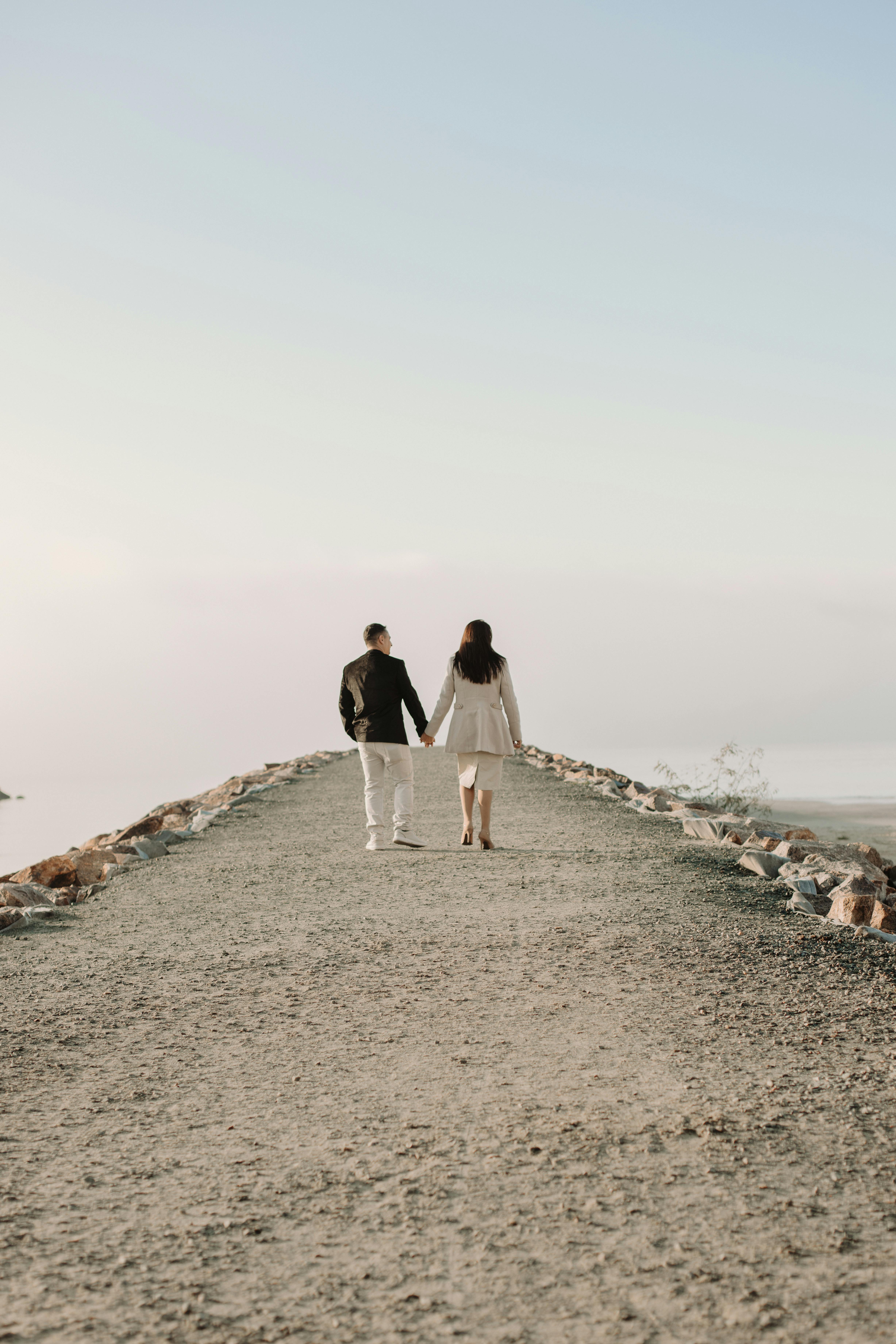 A couple holding hands walks on a scenic beach pathway during a serene day.