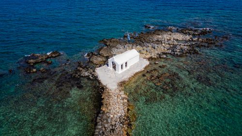 Free Aerial shot of a small white chapel on a rocky island surrounded by turquoise sea in Greece. Stock Photo