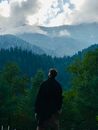 一名男子站在森林中，凝视着薄雾笼罩、阴云密布的山脉