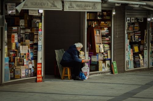 Free Elderly man reading at a sidewalk bookshop, surrounded by a variety of books. Stock Photo