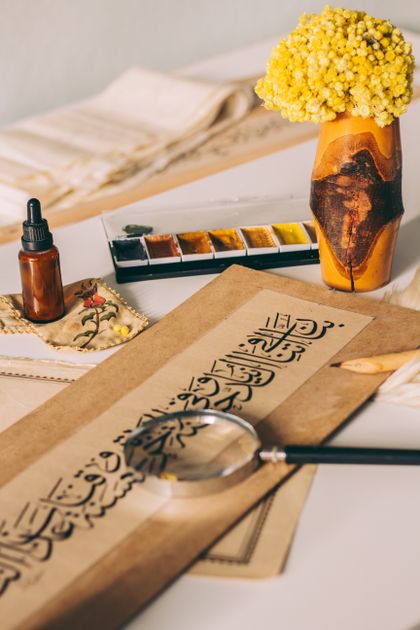 Elegant Arabic calligraphy artwork created with ink paints, surrounded by vibrant colours and fresh flowers on a studio table