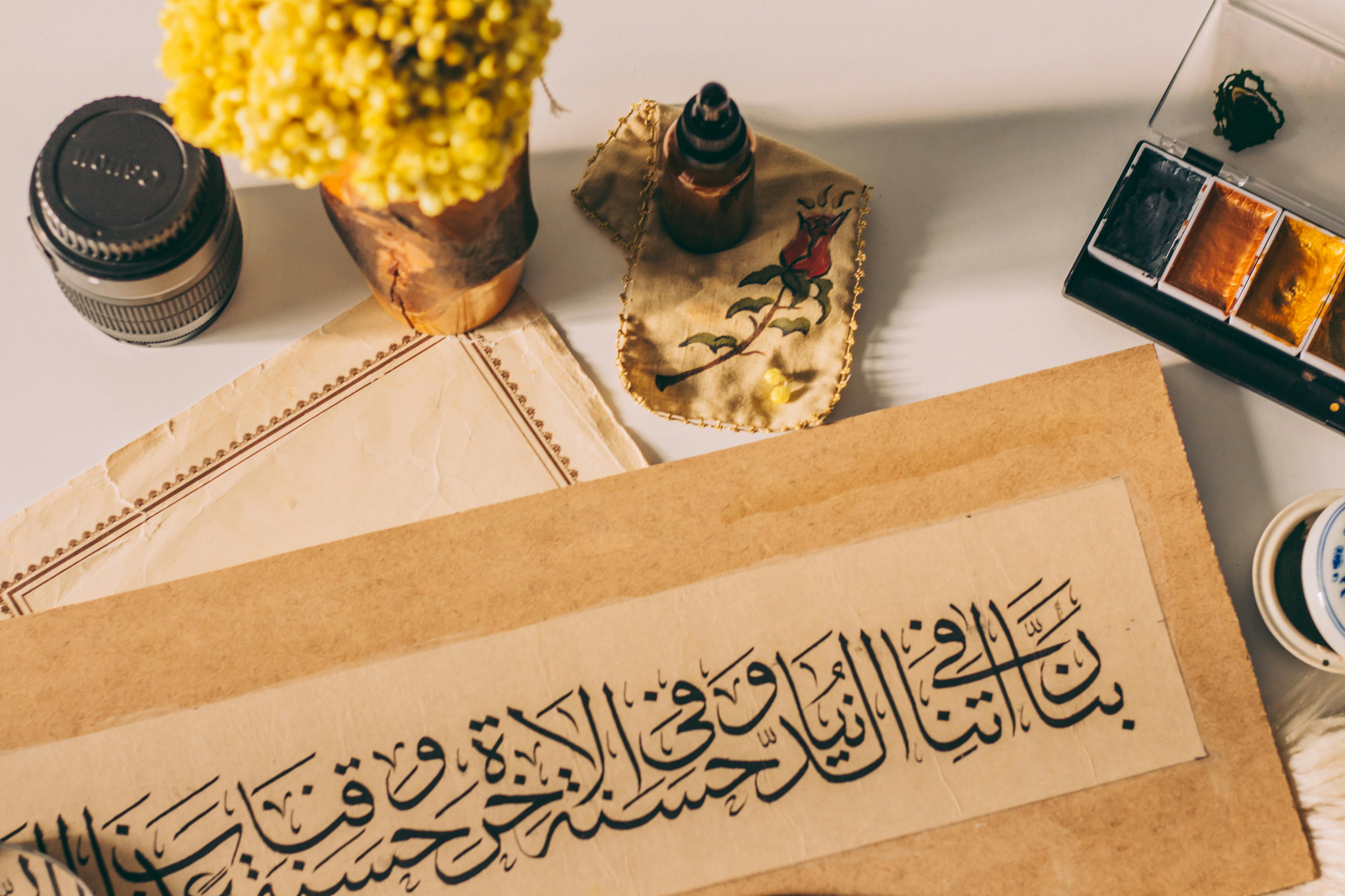 Artistic setup featuring Arabic calligraphy, flowers, ink, and watercolor paints on a desk.