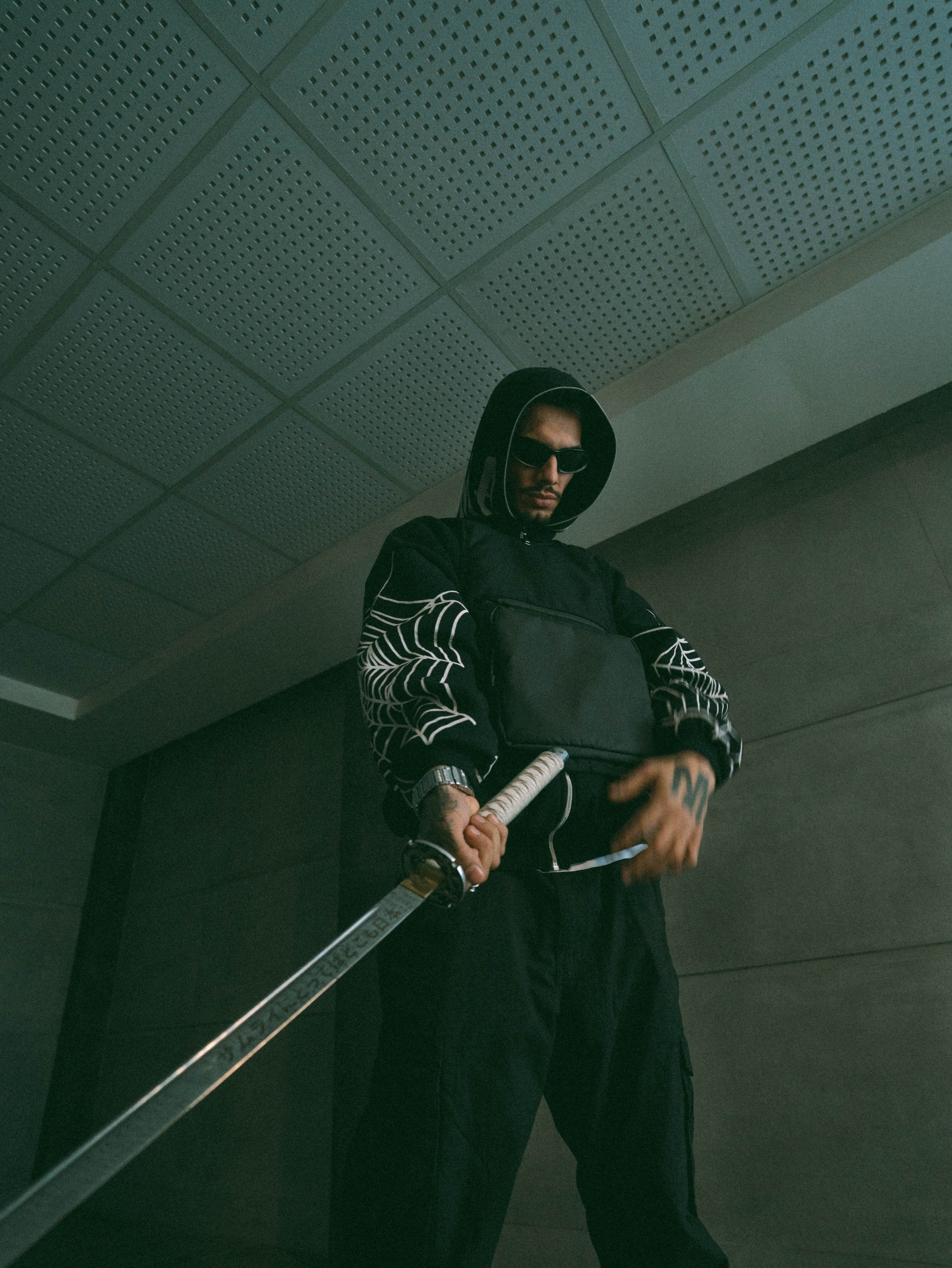 Man in dark hoodie poses with katana indoors, evoking a mysterious and dramatic urban vibe.