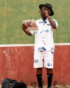 A fashionable man dressed in eclectic attire reading a newspaper outdoors against a vibrant backdrop.