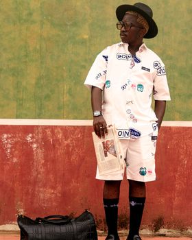 Fashionable man in graphic attire holding a newspaper, standing outdoors with luggage.