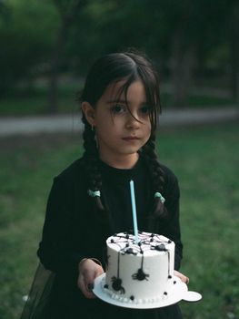 A young girl in black attire holds a Halloween-themed cake with spider decorations.