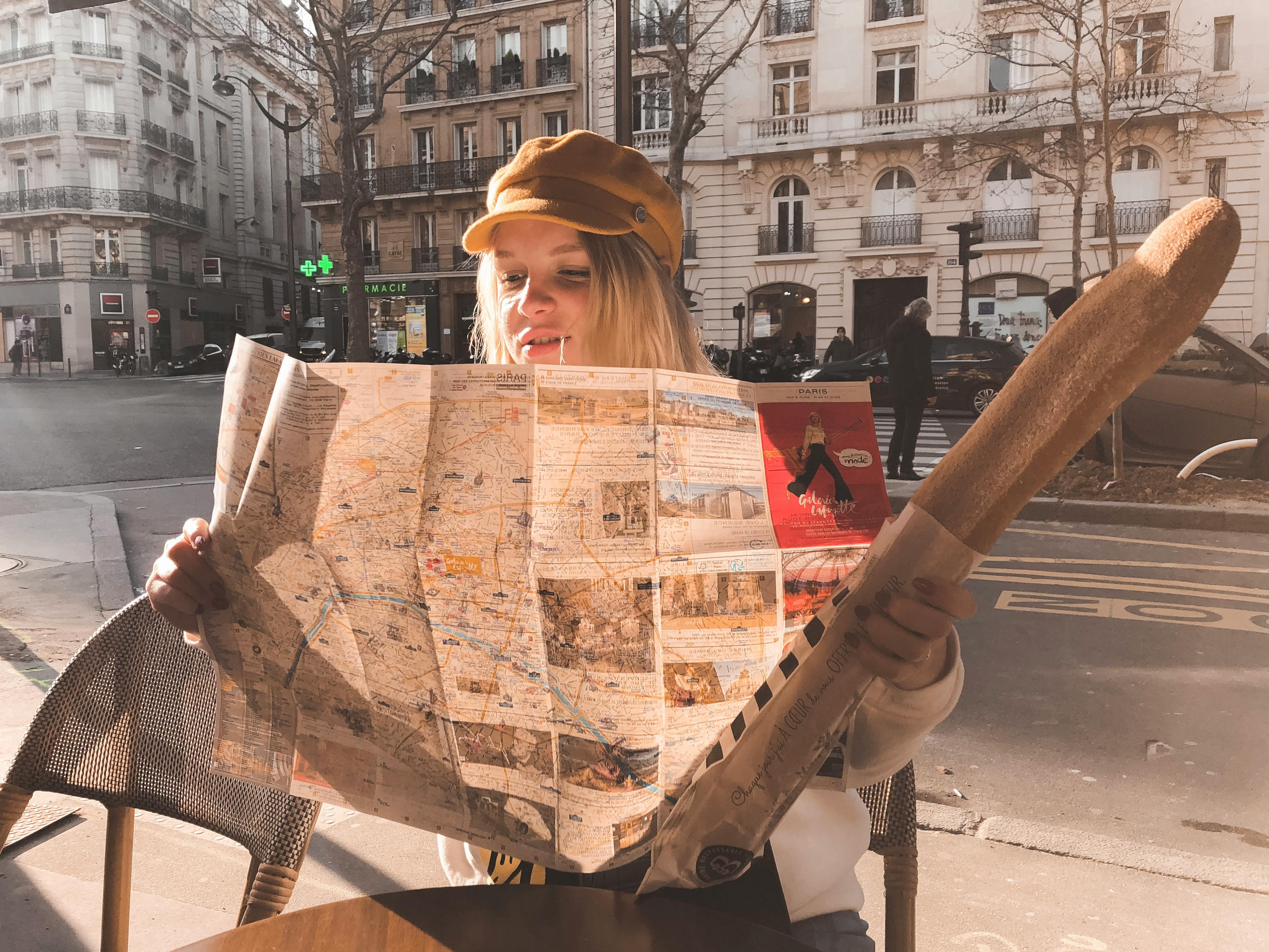Blonde woman reading a map and holding a baguette at a Parisian street cafe.