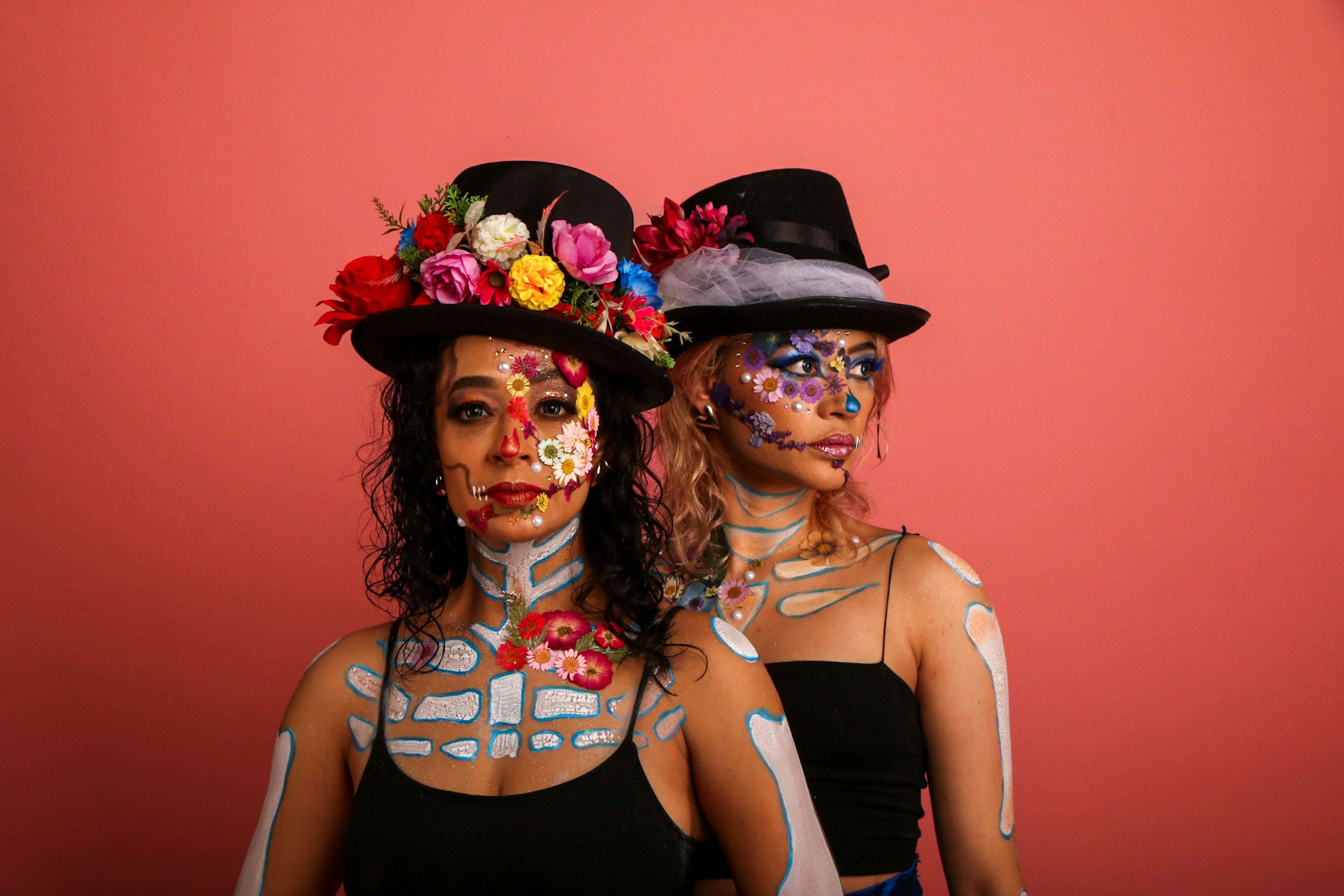 Two women with artistic face paint and floral hats against a vibrant backdrop.