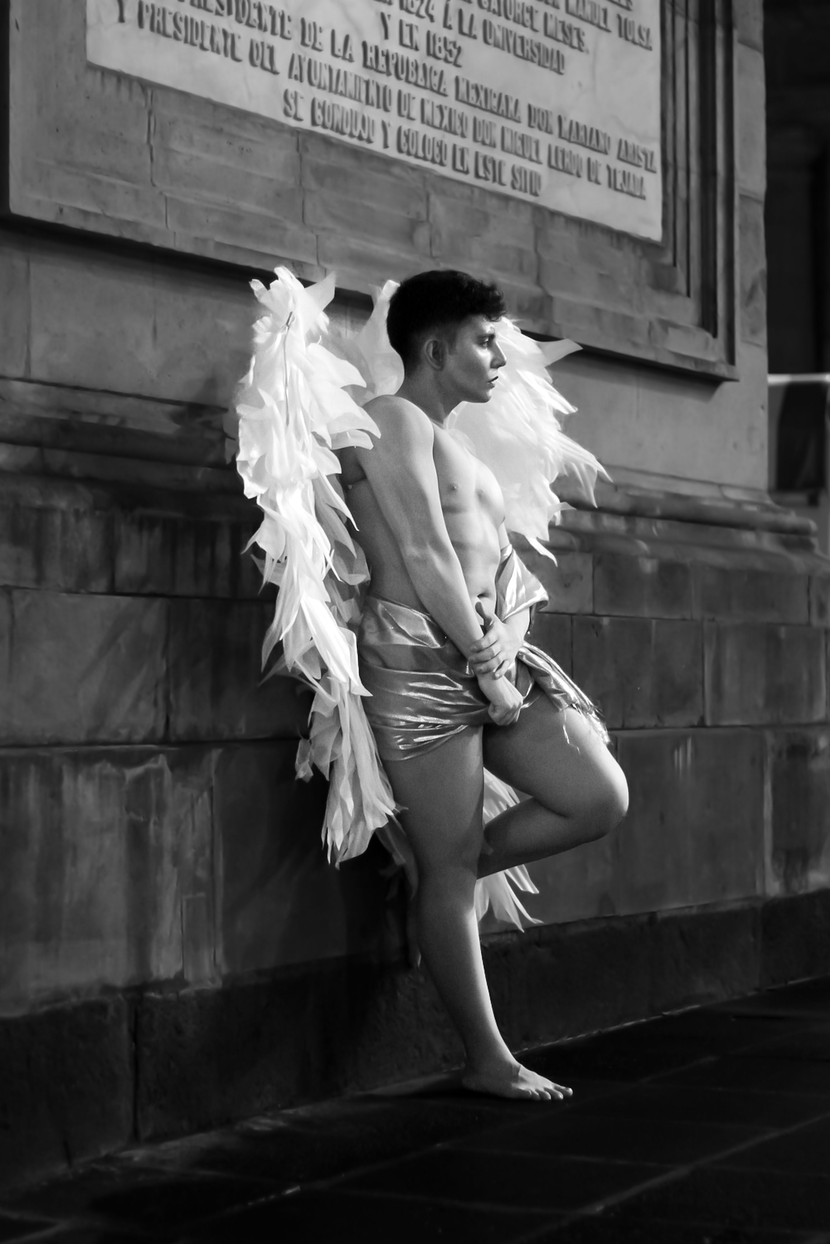 A person with angel wings poses dramatically against a stone wall in black and white.