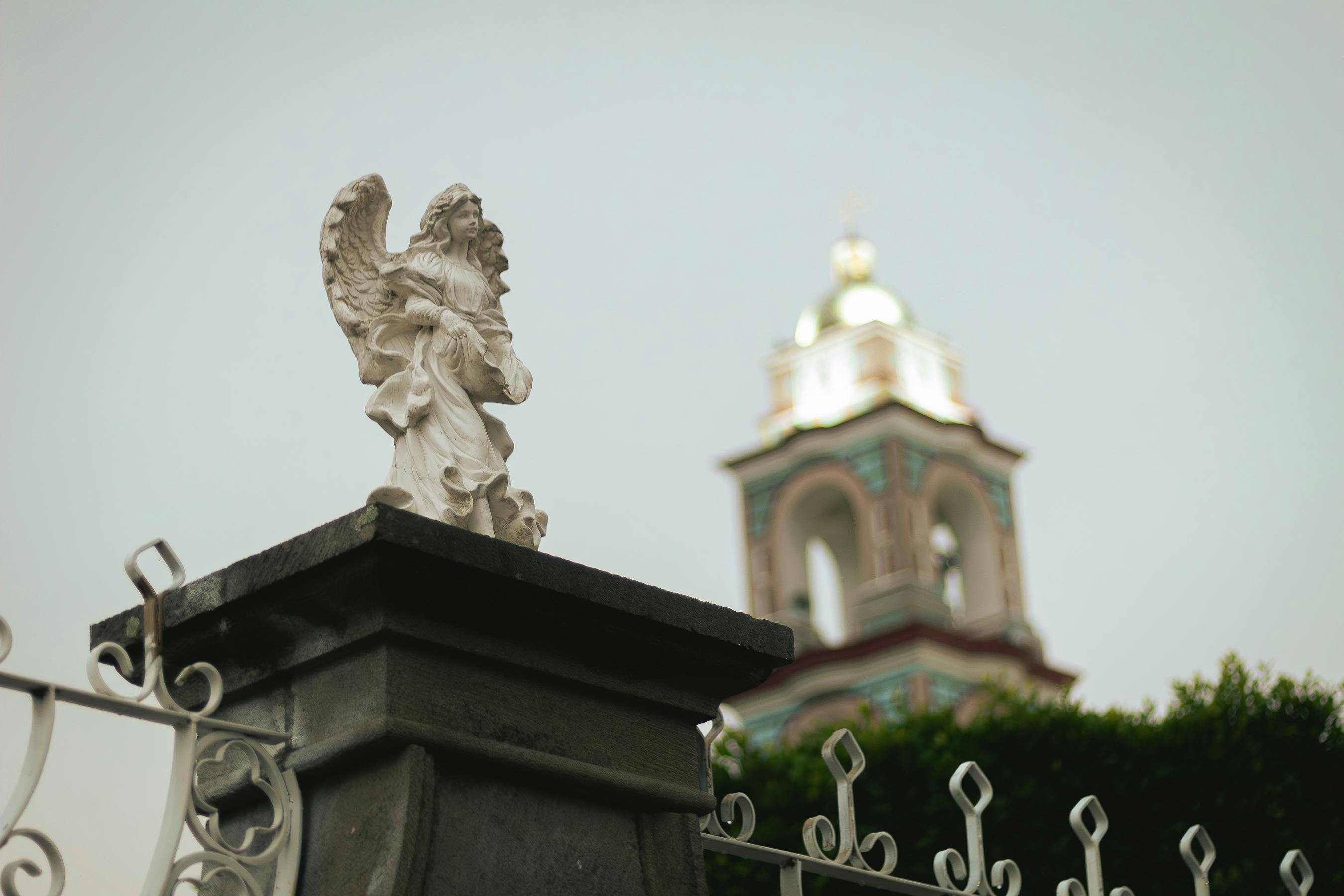 Angel statue atop stone pillar with blurred church spire in background, creating a serene scene.