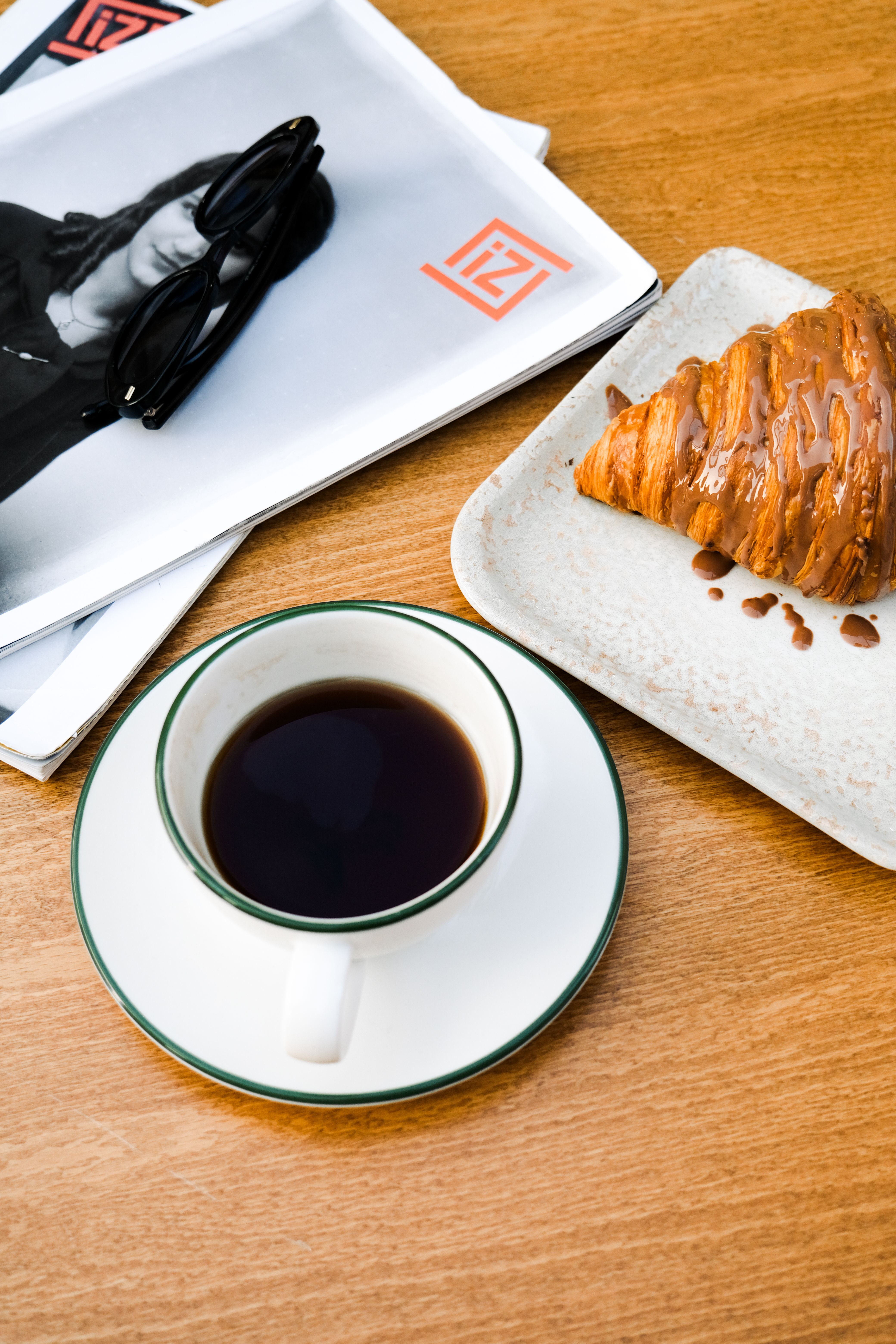 A cup of coffee with a croissant and magazines on a wooden table.