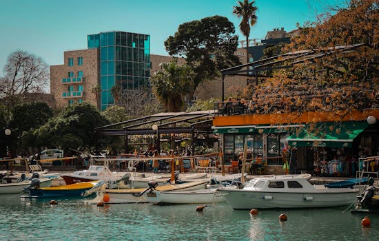 Vibrant marina in Budva with boats docked by bustling cafés, framed by lush greenery. by Ender Vatan