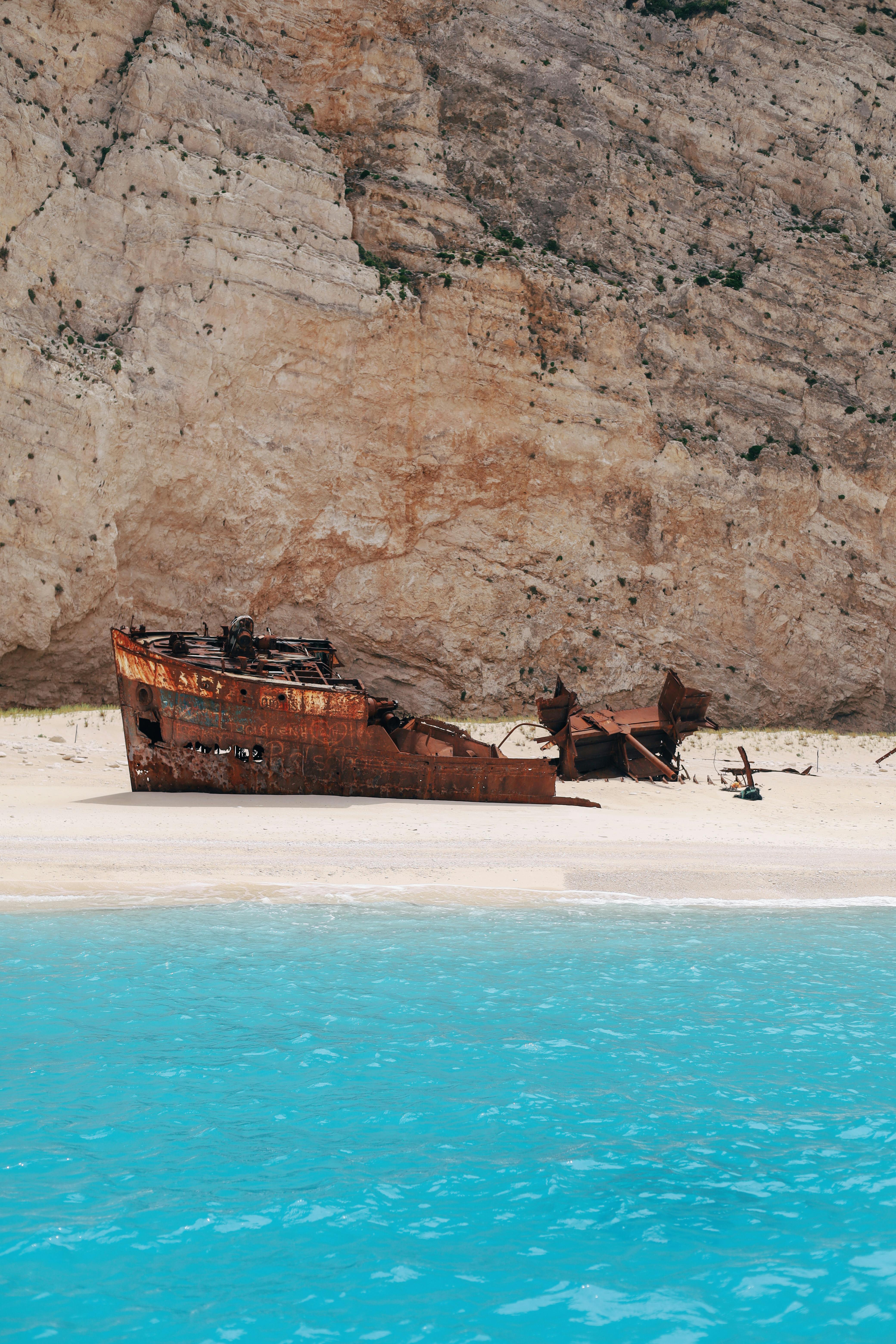 Abandoned shipwreck on a sandy beach against rocky cliffs, serene turquoise water.