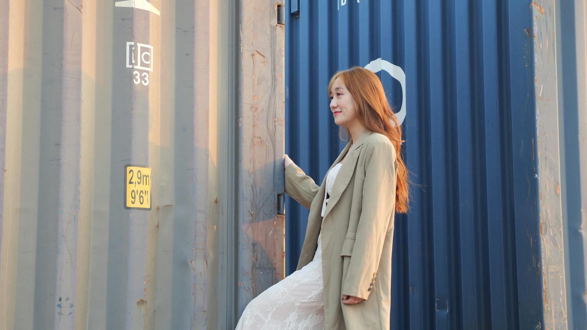 Young woman in coat posing against shipping containers outdoors in soft lighting.