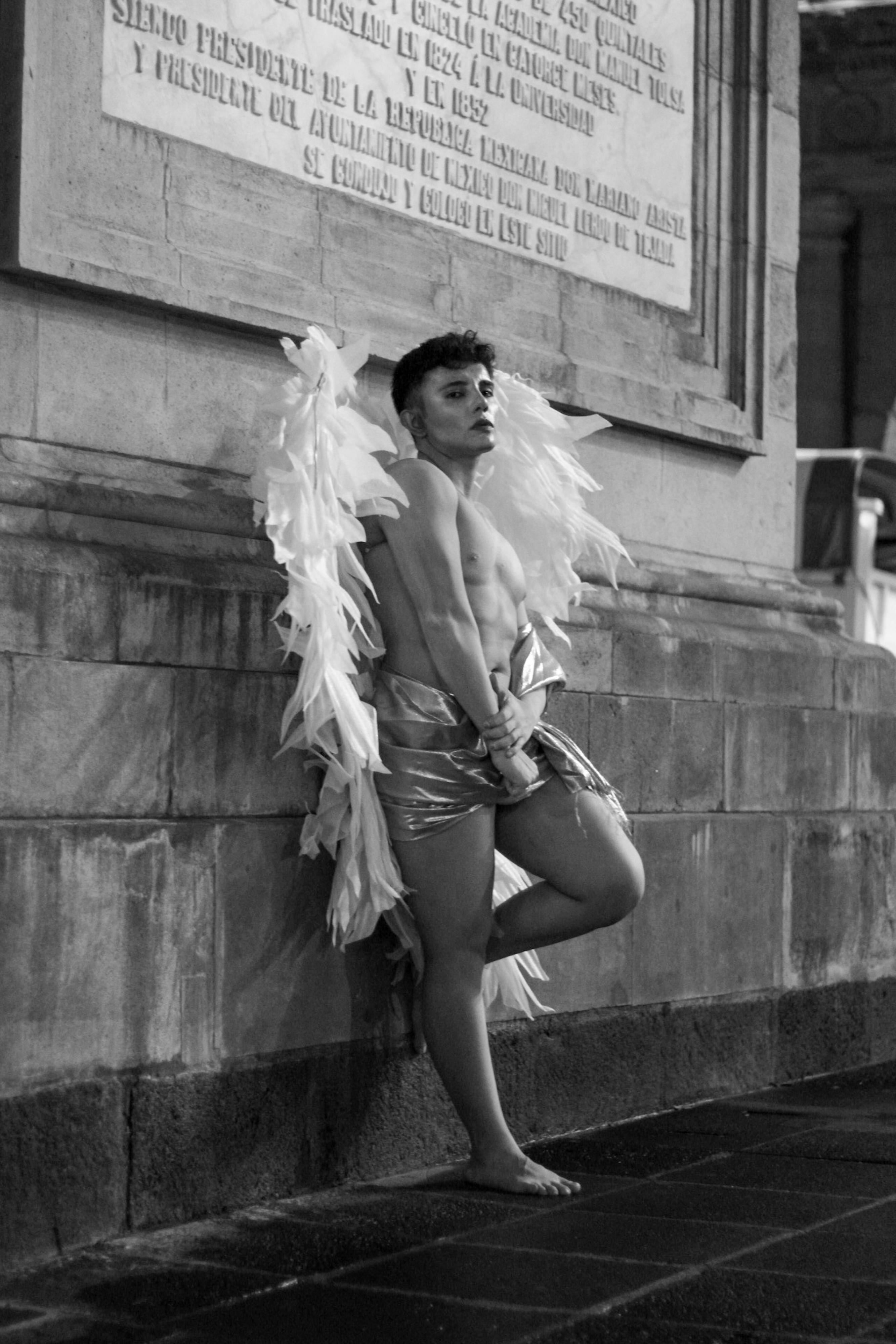Black and white photograph of a person in an angel costume against a historic wall.