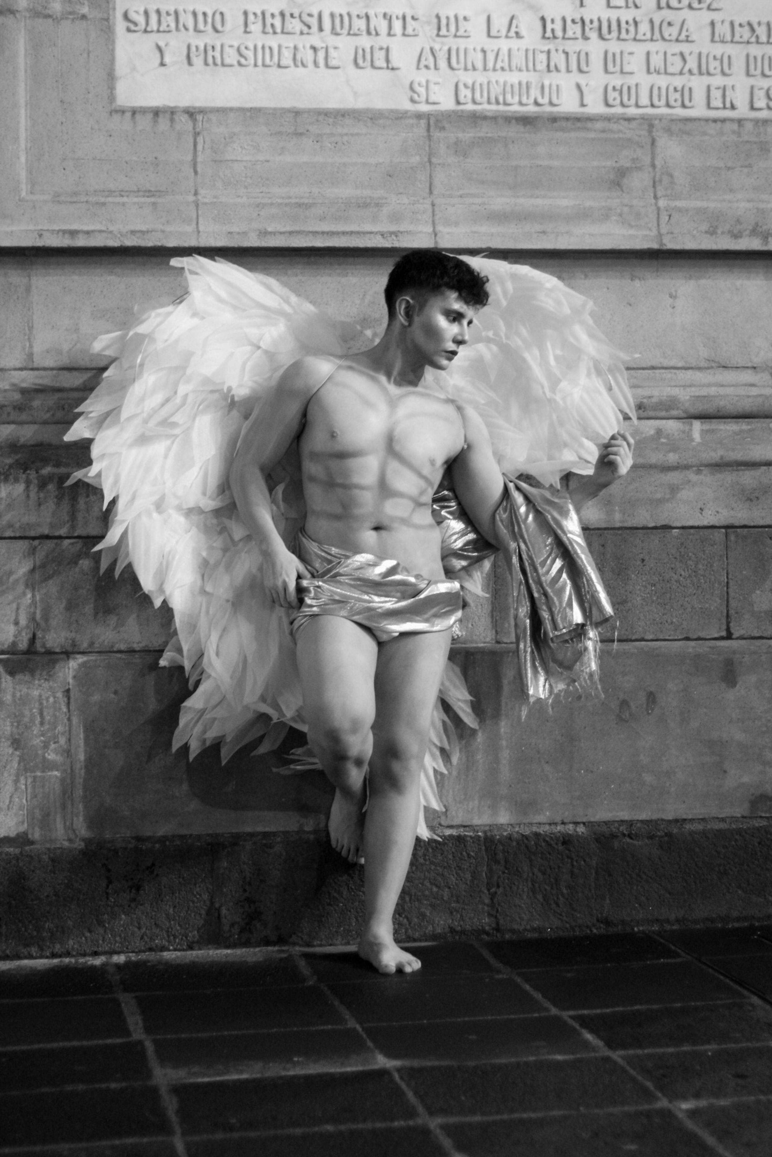 Black and white photo of a muscular man dressed as an angel leaning against a wall.