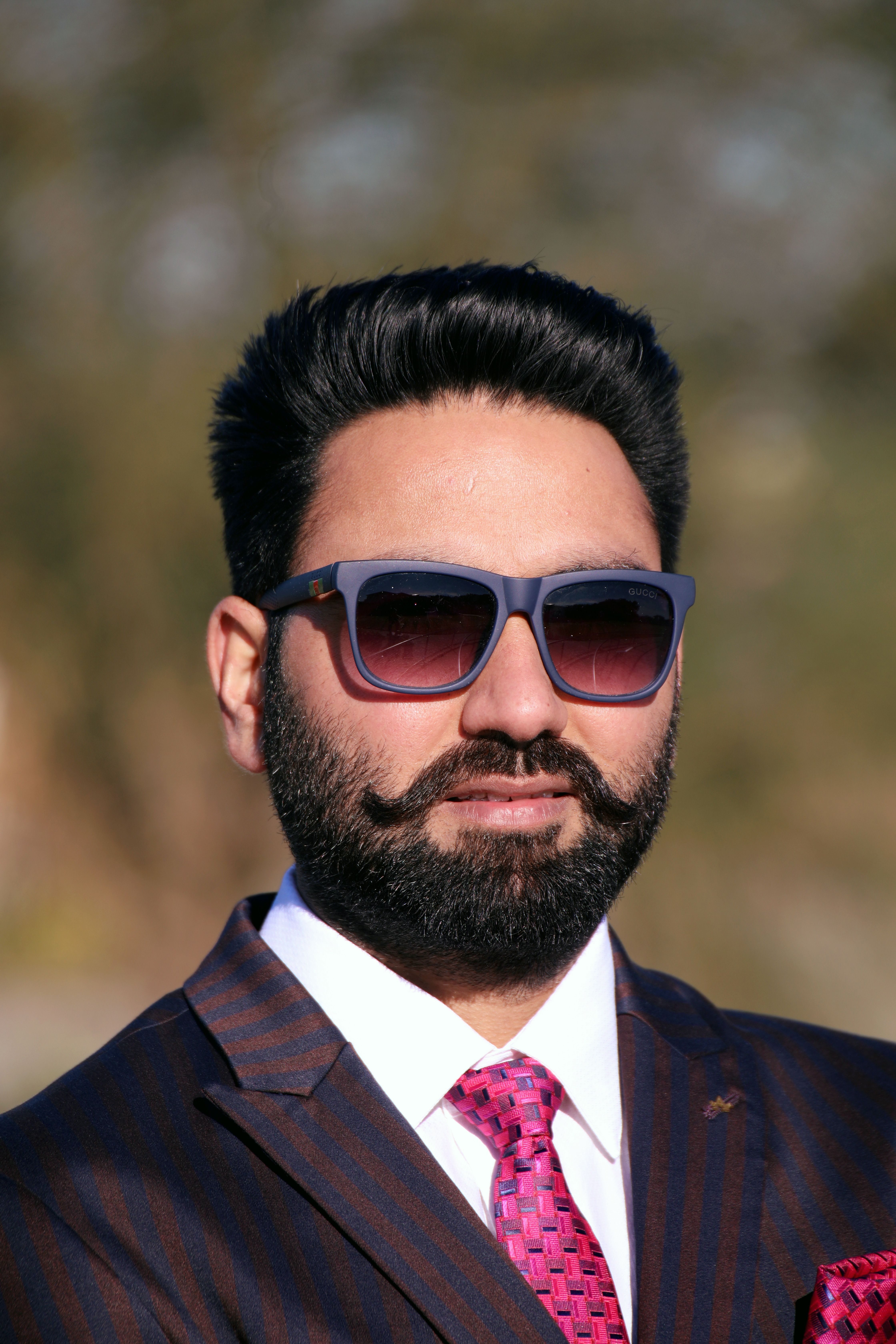 Portrait of a fashionable man with sunglasses and a beard, wearing a suit outdoors.