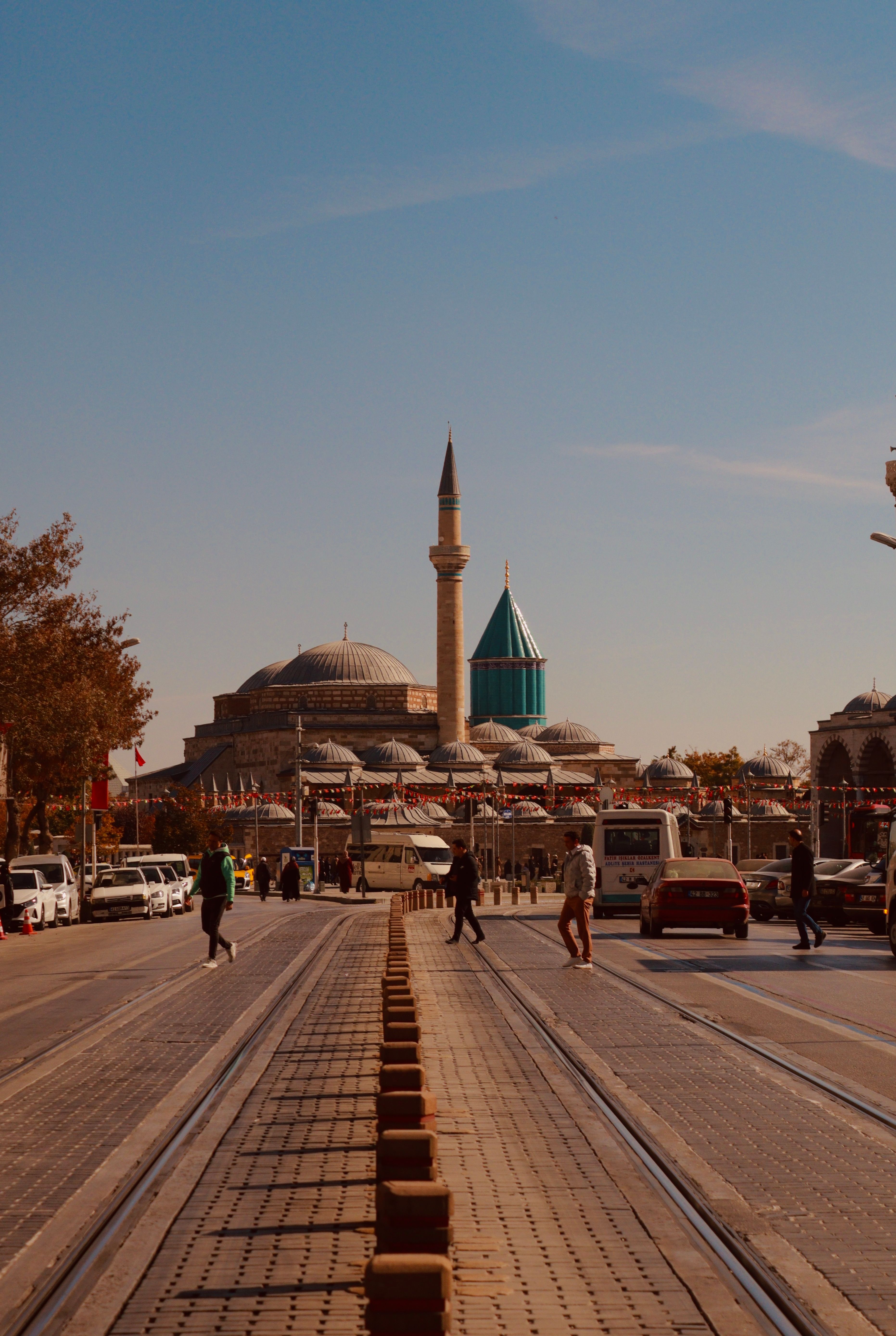 A street view of the iconic Mevlana Museum in Konya, Türkiye, with people and vehicles.